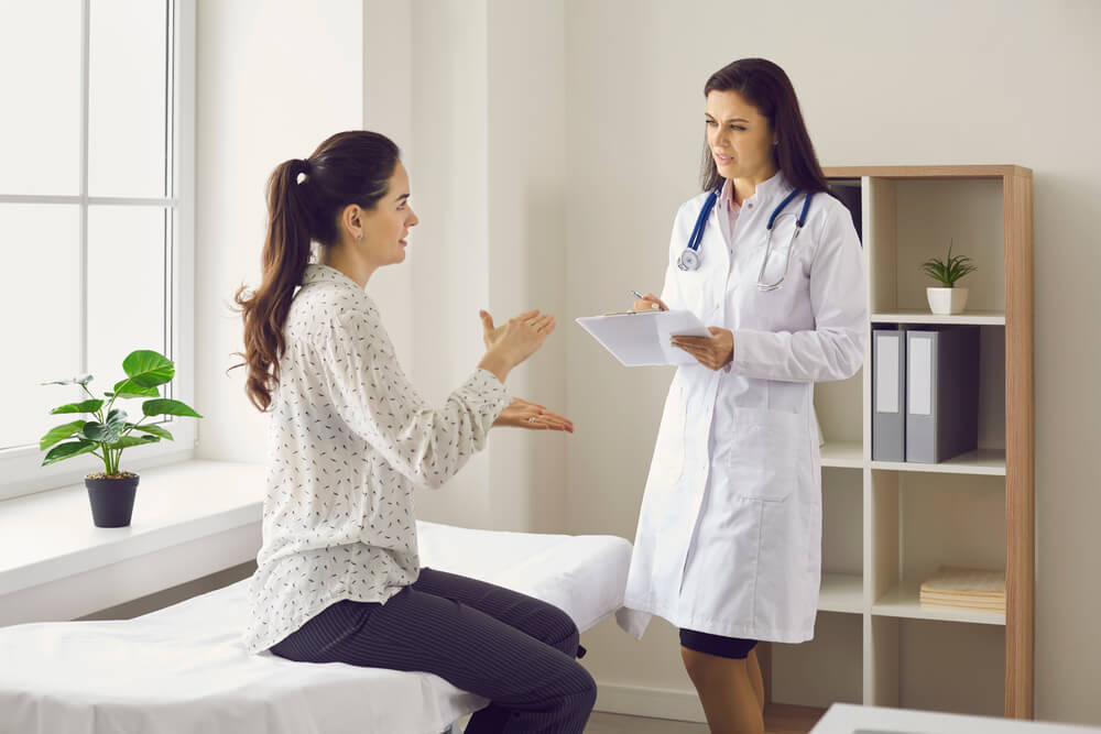 Woman Talking To Female Doctor Sitting on Examination Bed at Clinic.