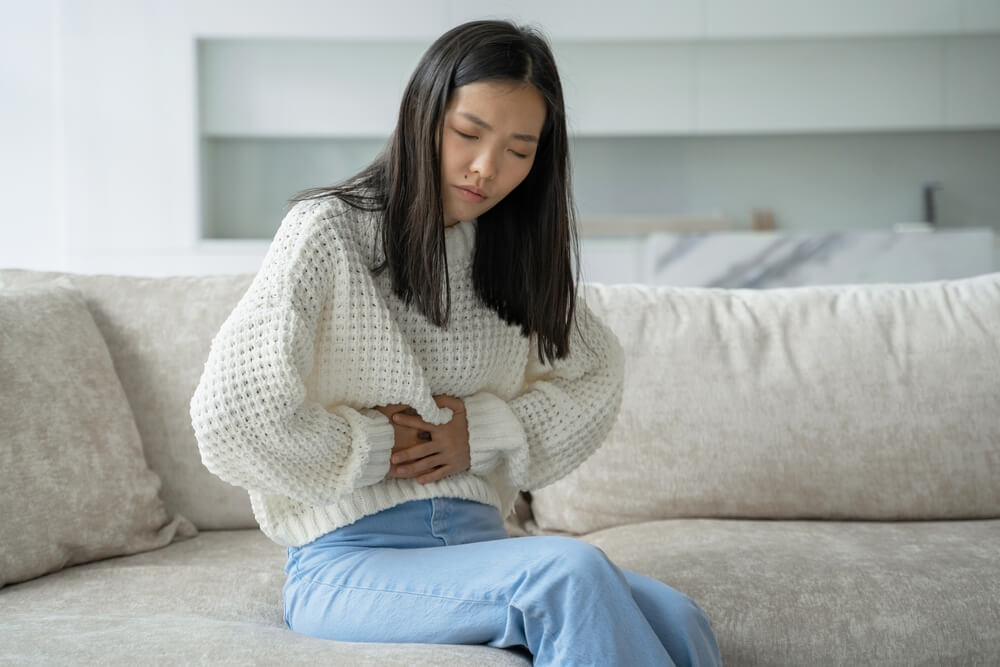 An Asian Woman in a White Blouse Is Sitting on the Couch and Feels a Strong Abdominal Pain