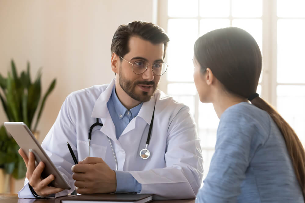 Smiling Young Caucasian Male Doctor in White Medical Uniform Consult Female Patient Using Tablet on Consultation