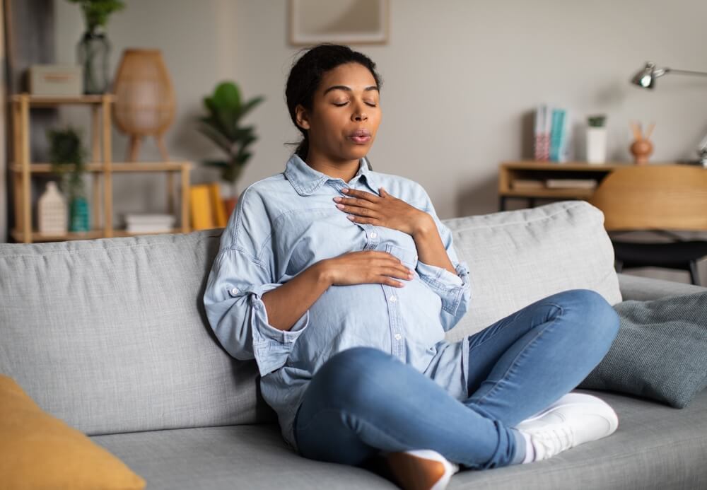 Pregnancy Yoga. Pregnant African American Woman Doing Breathing Exercise With Eyes Closed Relaxing Sitting on Couch at Home