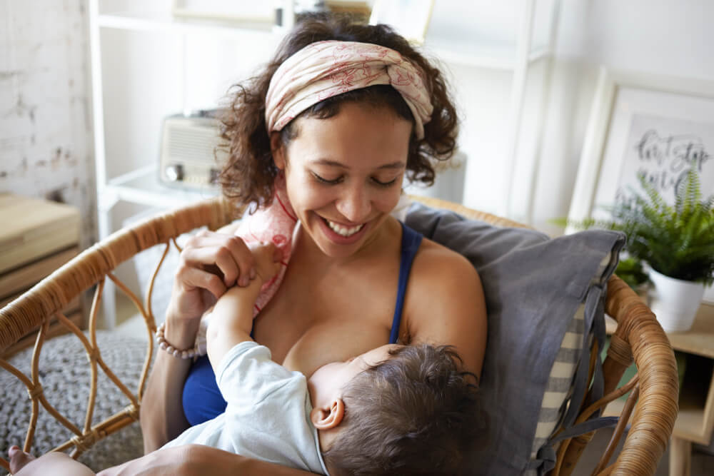 Motherhood, Infantry, Parenting And Childhood Concept. Picture Of Cheerful Young Brunette Hispanic Woman Wearing Headscarf Smiling Broadly, Nursing Her Infant