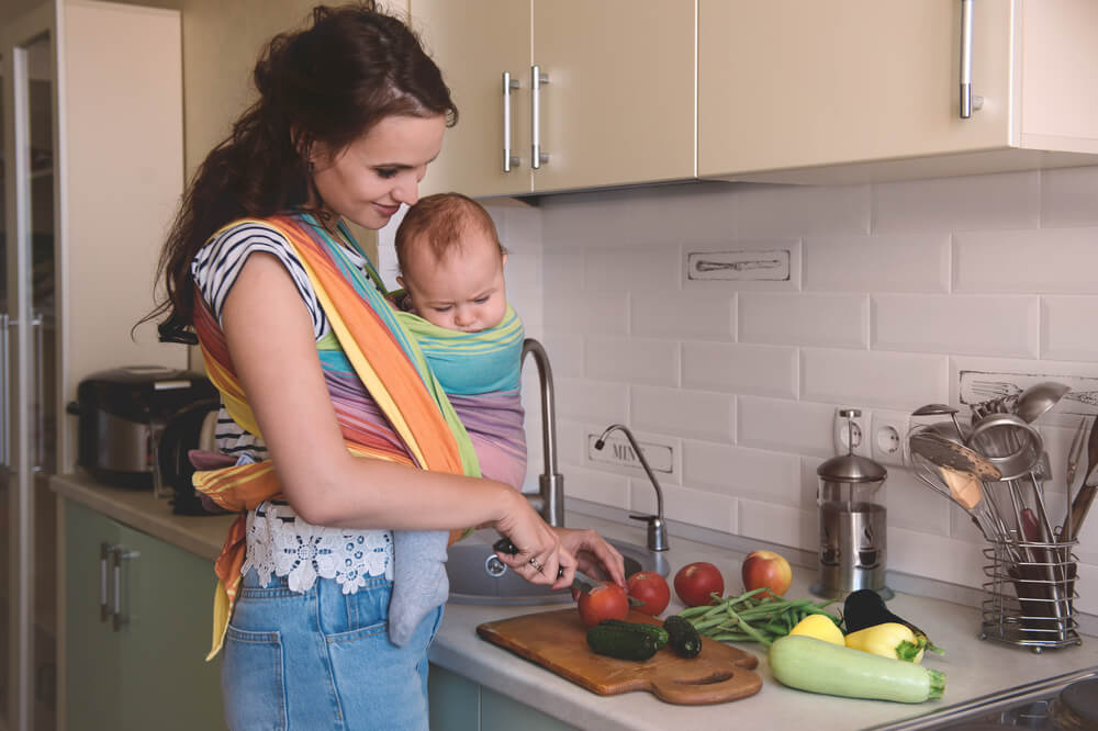 Young Mom Cooking In The Kitchen With The Little Baby In A Sling. Vegetarian Healthy Food. Healthy Food Breastfeeding Mothers.