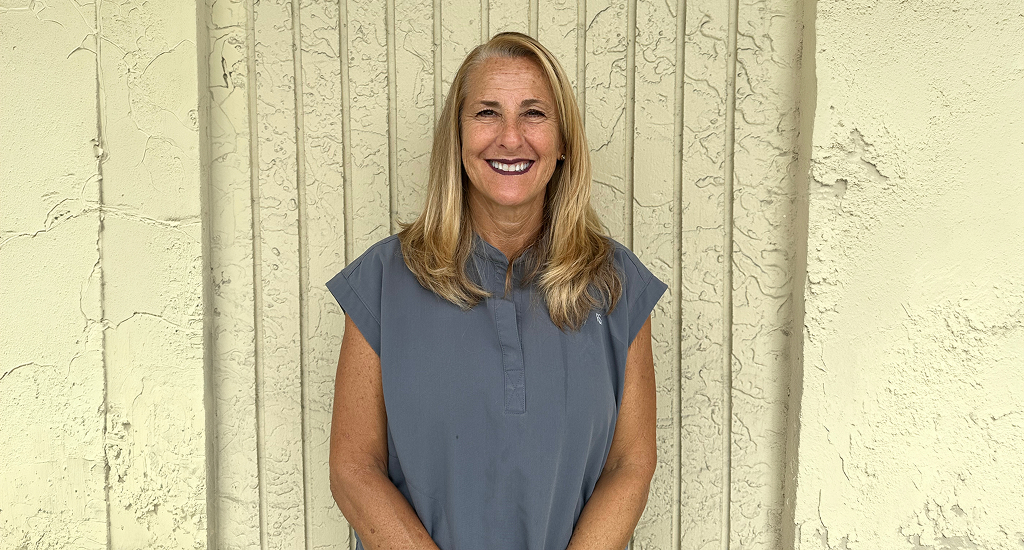2045a52075acfa61362960841c631d6c25d000a4 1 Portrait of a smiling woman with blond hair in a blue-gray blouse, standing against a beige textured wall.