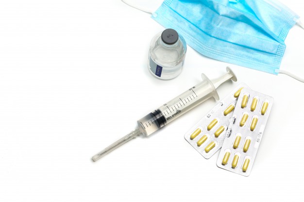 Face mask, Pills, Vaccine Dose and a Syringe on a white Surface