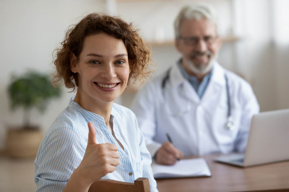 Smiling Female Patient Sitting at Doctor’s Office Showing Thumbs Up