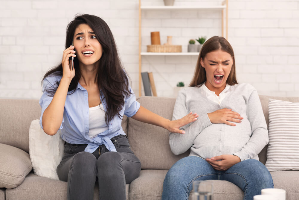 Girl Calling Doctor for Her Pregnant Friend Having Spasm Suffering From Pain Sitting on Sofa Indoor.