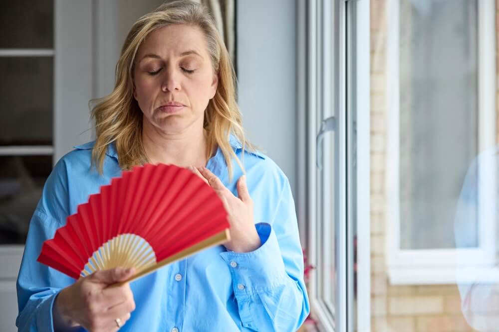 Menopausal Mature Woman Having Hot Flush At Home Cooling Herself With Handheld Paper Fan
