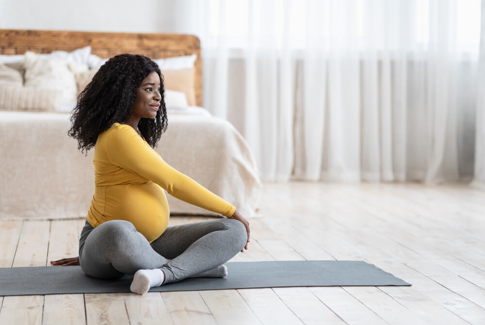 Sporty Black Pregnant Woman Sitting on Yoga Mat, Exercising at Bedroom