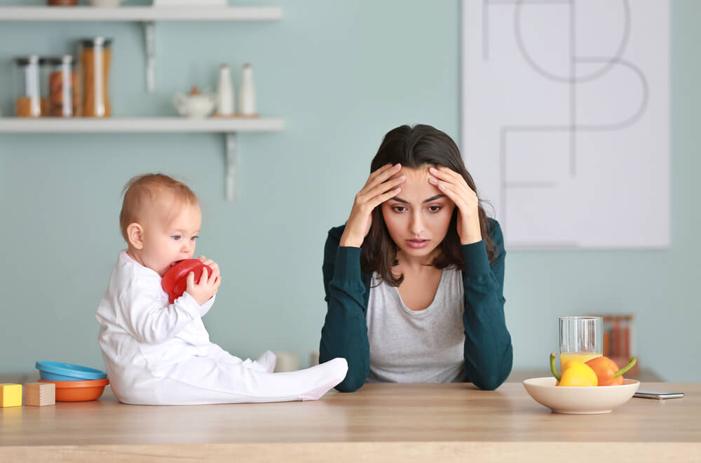 Young Woman Suffering From Postpartum Depression at Home With Her Baby Sitting on the Table Next to Her