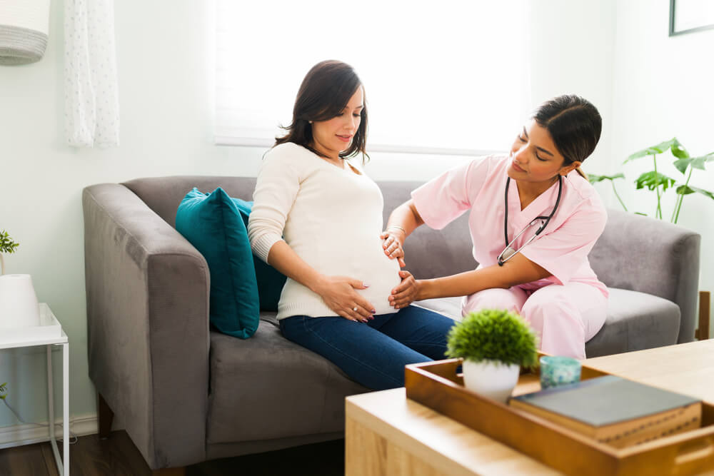 Hispanic Midwife In A Nursing Uniform Examining And Feeling The Baby In The Belly Of A Happy Pregnant Woman During A Home Visit