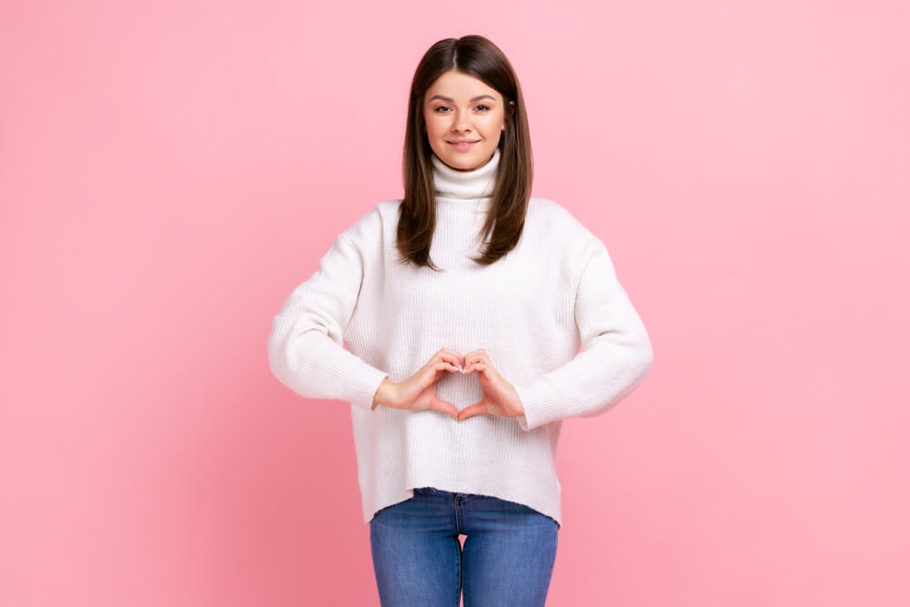 Positive optimistic brunette female showing heart shape in front of belly as symbol of women health, wearing white casual style sweater
