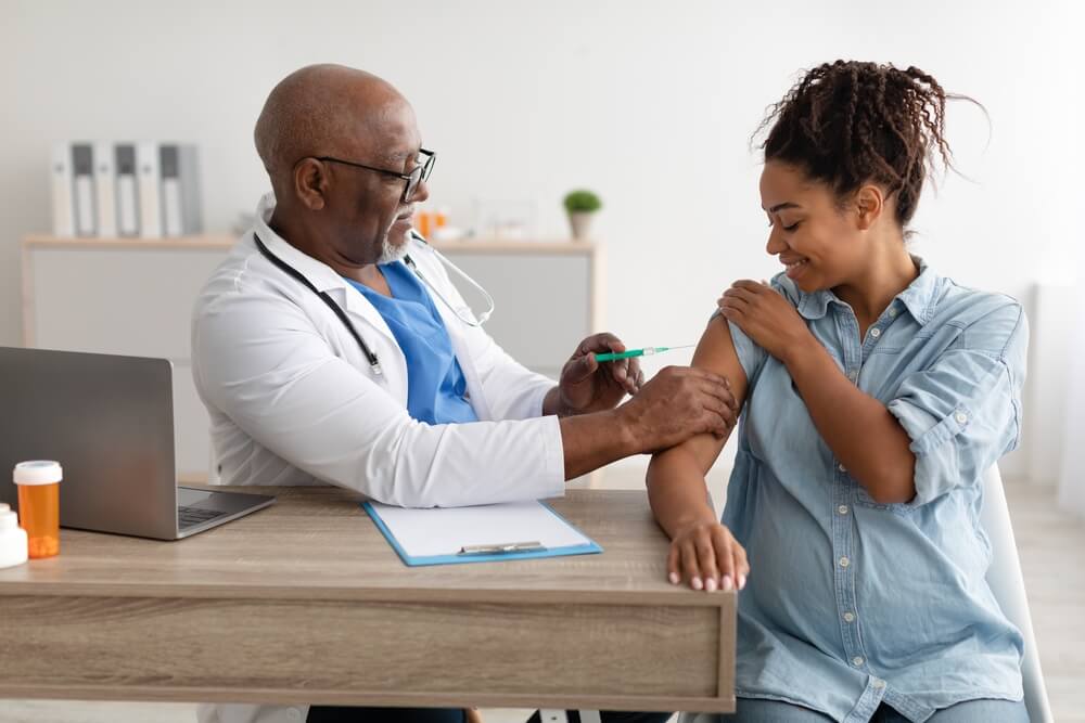 Smiling Pregnant Young Woman Getting Vaccinated At Clinic, Mature Male Doctor In Glasses Giving Vaccine Shot Injecting Mother-To-Be In Arm