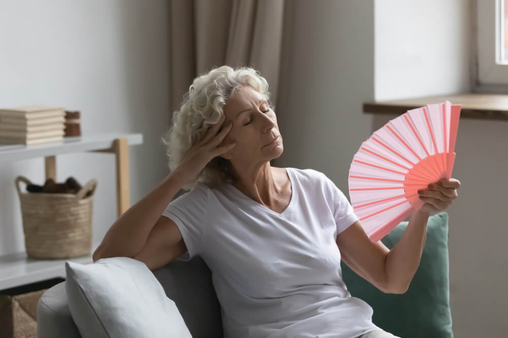 Overheated Senior Woman Relax Sit On Sofa Waving Orange Peach Colour Fan Cool Herself, Feels Unwell Due Unbearable Hot Weather, Discomfort and Hormonal Changes