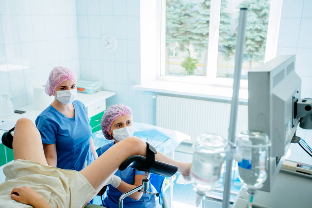 Female Gynecologist and Assistant in Mask and Hair Cap Working Unrecognizable Patient in Gynecology Chair