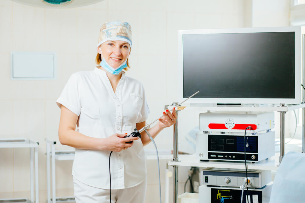 Portrait of Happy Female Surgeon in Protective Wear Uniform Holding Hysteroscope for Laparoscopy Operation in Empty Operating Room.