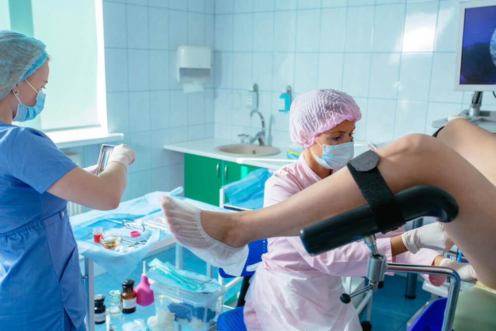 Female Gynecologist With Young Woman Assistant in Mask and Hair Cap With Unrecognizable Patient in Gynecology Chair.