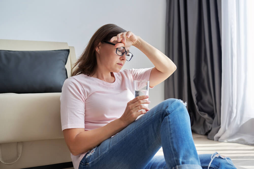 Tired overworked sad mature woman with glass of water sitting at home on the floor. Midlife crisis, health problems, negative emotions, mental health