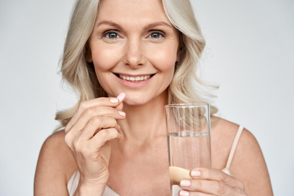 Smiling Happy Healthy Middle Aged 50S Woman Holding Glass of Water Taking Dietary Supplement Vitamin Pink Pill Isolated on White Background