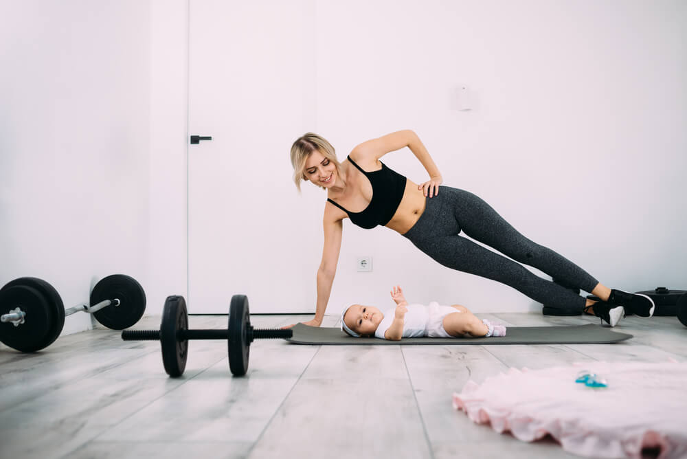A Blonde Mother Is Doing Fitness With Her Little Baby on the Floor in the Room.