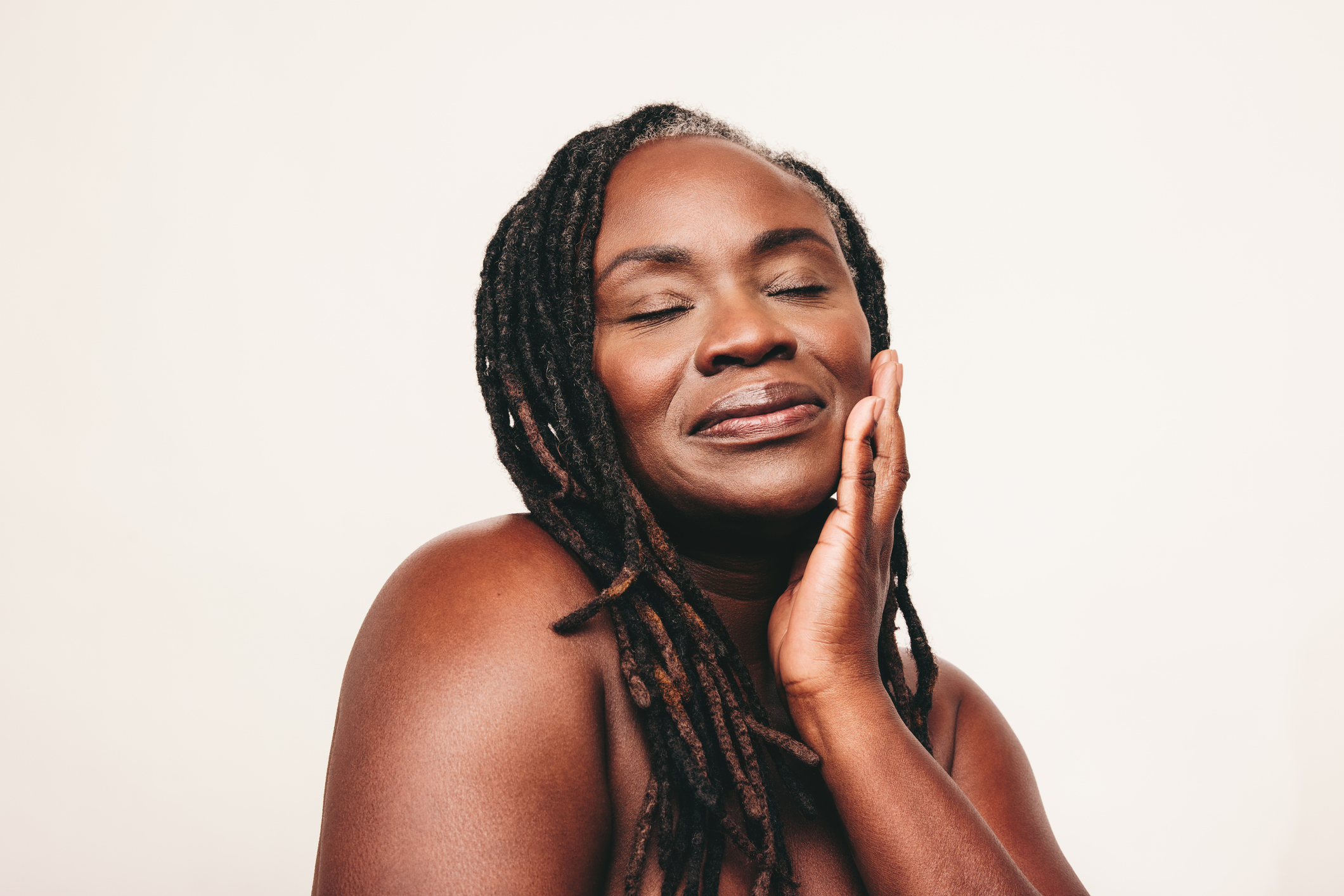 Beautiful woman with dreadlocks touching her flawless skin with her eyes closed. Confident dark-skinned woman embracing her smooth melanated skin. Mature black woman ageing gracefully.