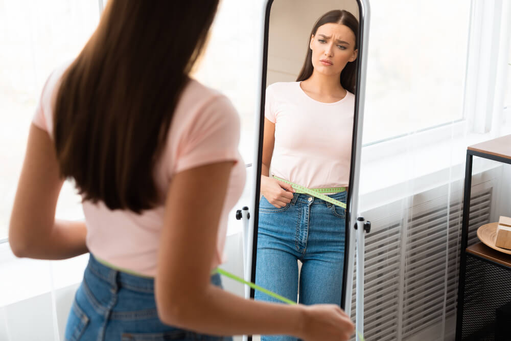 Sad Woman Measuring Waist With Tape After Weight Gain Standing in Front of Mirror at Home.