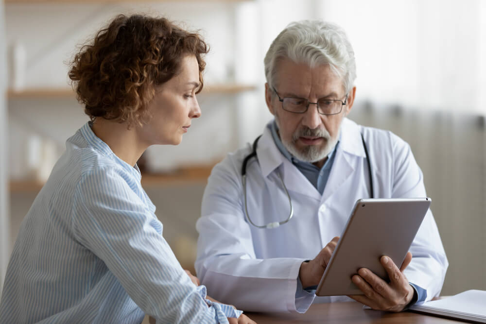 Older Adult Doctor Consulting Young Female Patient Using Digital Tablet Tech During Checkup Appointment Meeting. 