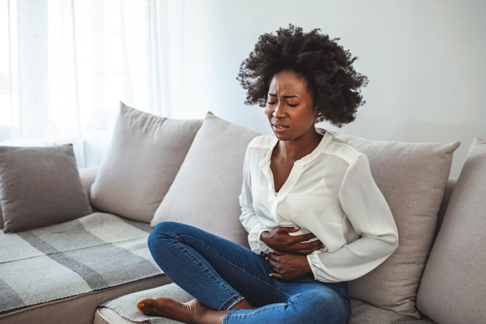 Woman Sitting On Sofa Suffering From Stomach Pain.