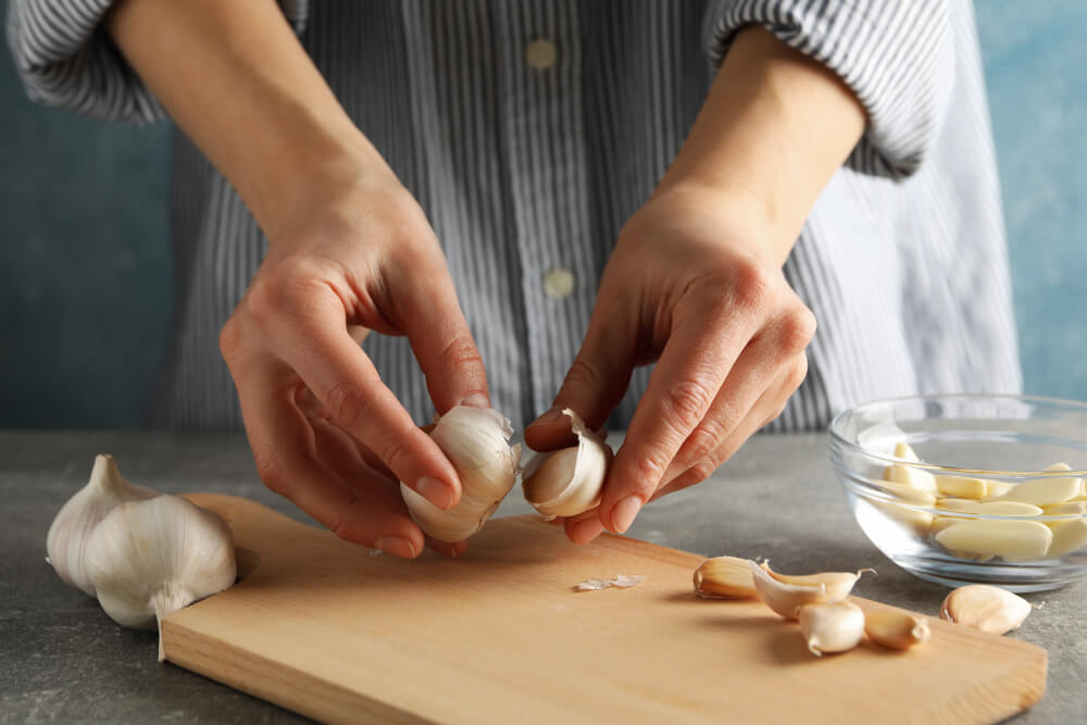 Women With Garlic Bulbs in Her Hands, Bowl of Garlic Slices, Board on Grey Wooden Background.