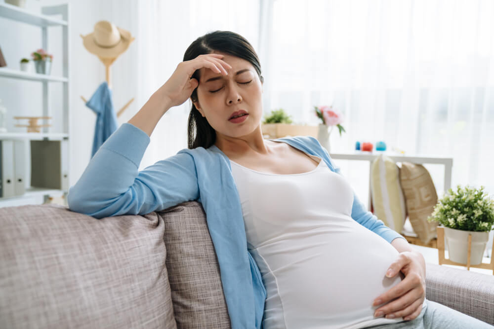 Young Asian Chinese Pregnant Woman With Headache Sitting on Sofa Couch in Living Room