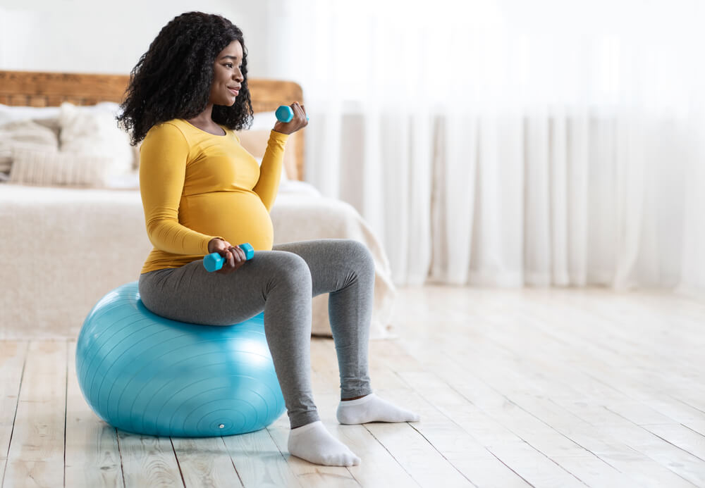 Active pregnant black lady exercising with barbells while sitting on fitness ball and looking at copy space, bedroom interior. 