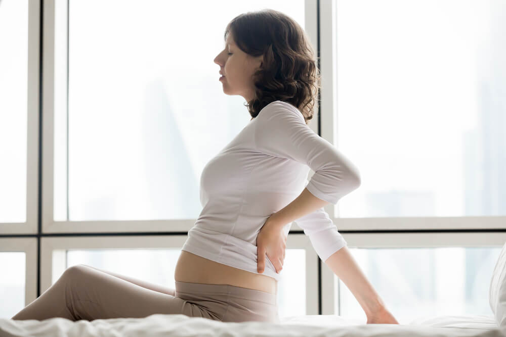 Healthcare, treatment. Side view portrait of young attractive pregnant woman sitting indoor on the bed touching aching back.