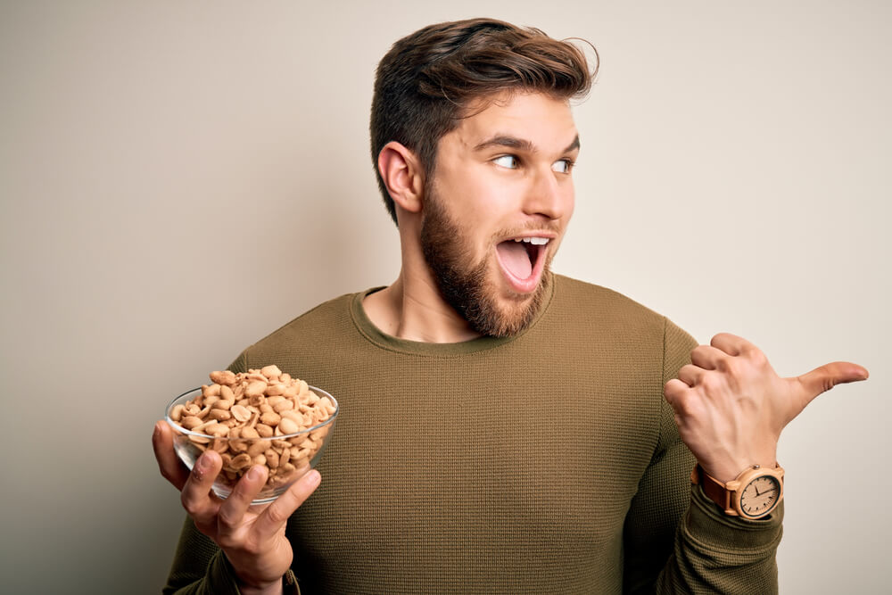 Young Blond Man With Beard and Blue Eyes Holding Bowl With Healthy Salty Peanuts Pointing and Showing With Thumb up to the Side 