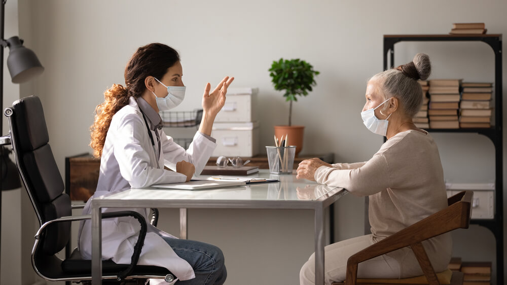 Side View Female Doctor Wearing Face Mask and Uniform Consulting Mature Patient About Treatment at Meeting in Hospital Office
