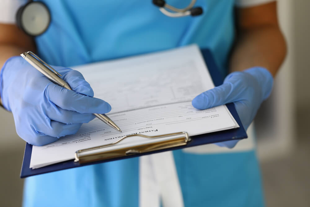 A doctor's gloved hands hold a clipboard with a pen and a patient registration document.