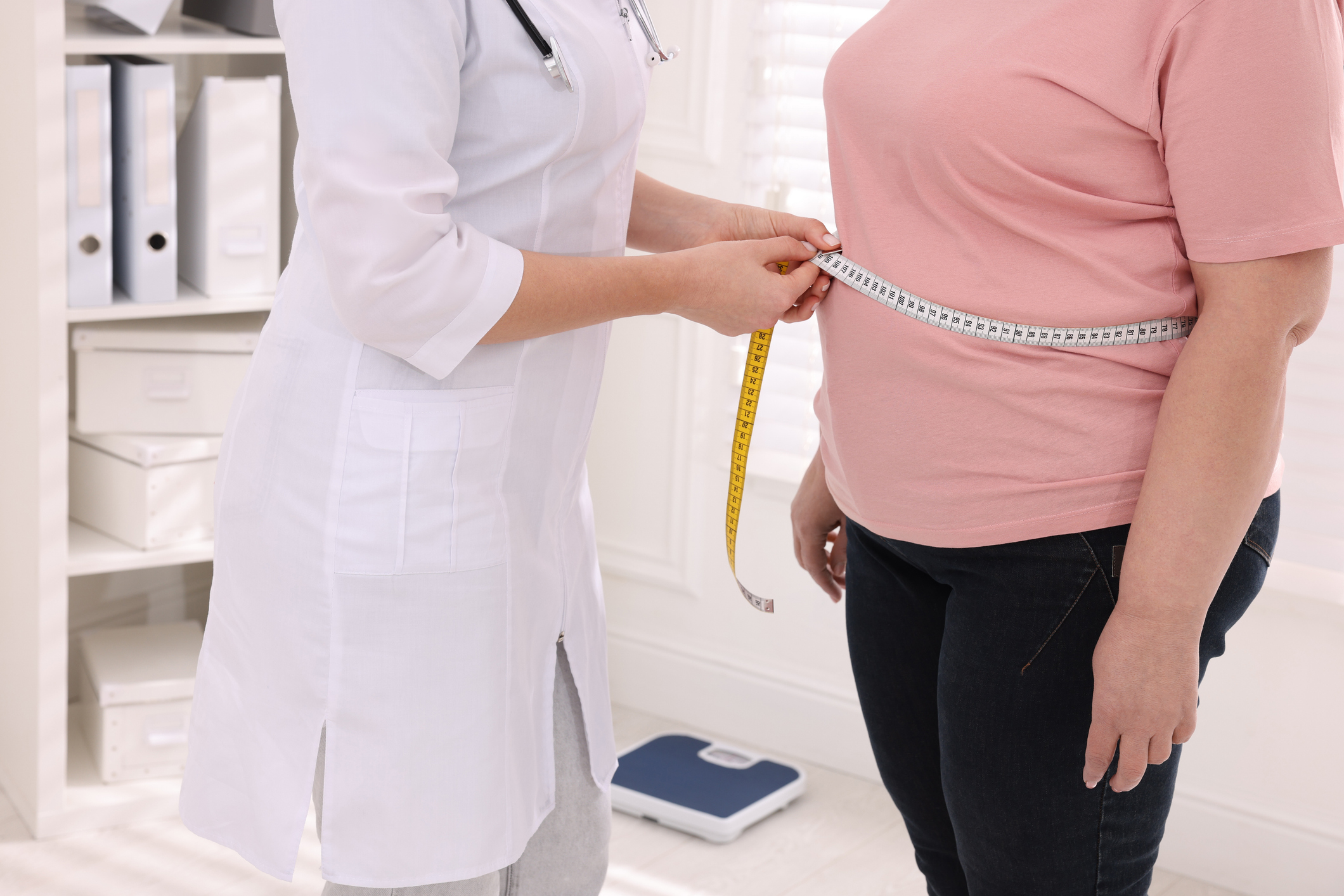 Nutritionist measuring overweight woman’s waist with tape in clinic, closeup