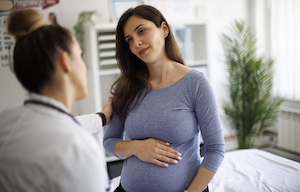 top_blog_non-stress-testing-nst_300x192 A pregnant woman stands beside an exam bed while speaking with her doctor during a prenatal appointment. The physician, seen in the foreground, gently touches the patient’s shoulder in a supportive gesture as the expectant mother rests a hand on her belly in a bright clinic room.