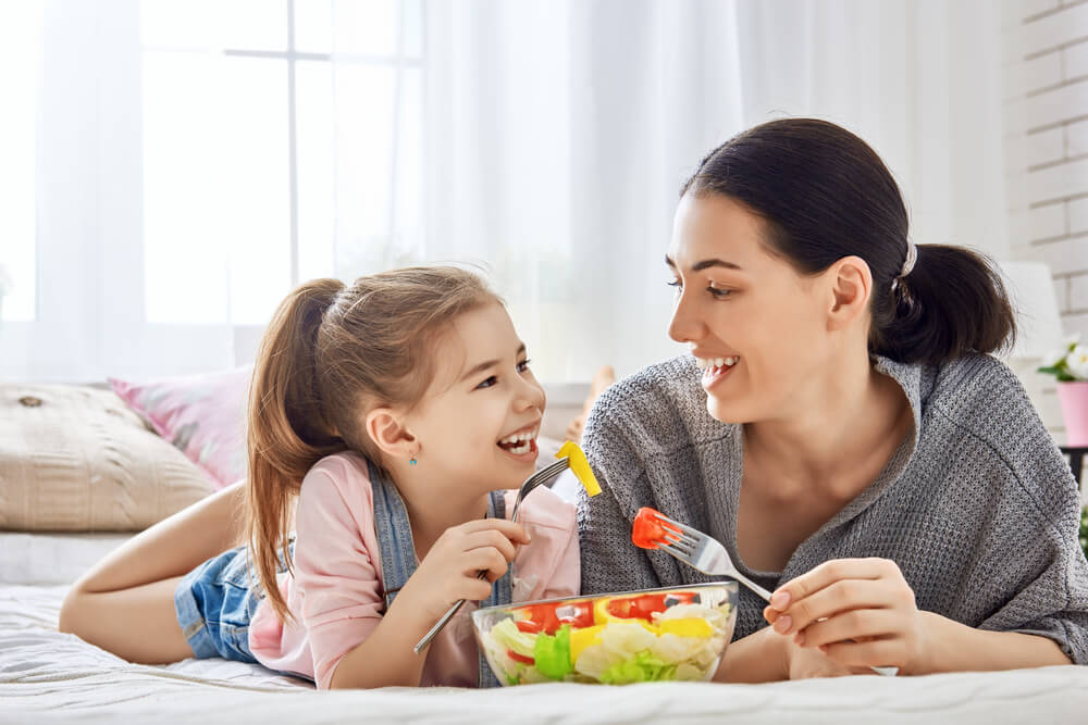 Mother and Her Daughter Child Girl Are Eating Salad on the Bed in the Room
