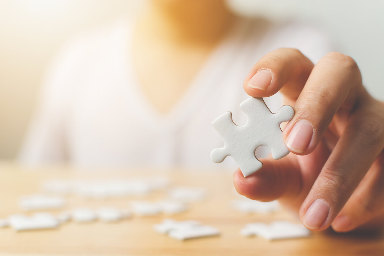 Hand of Male Trying to Connect Pieces of White Jigsaw Puzzle on Wooden Table.
