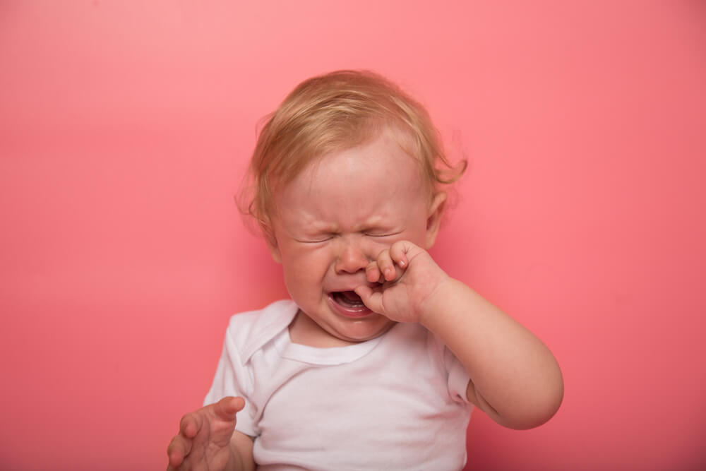 Beautiful Curly Blond Baby Girl Crying on a Pink Background With Finger in Mouth