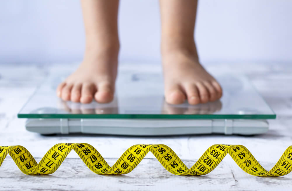 Weight Control Concept With Centimeter in Focus and Blurred Kid’s Feet on Digital Scale in the Background. Child Measuring Weight, Healthy Growing