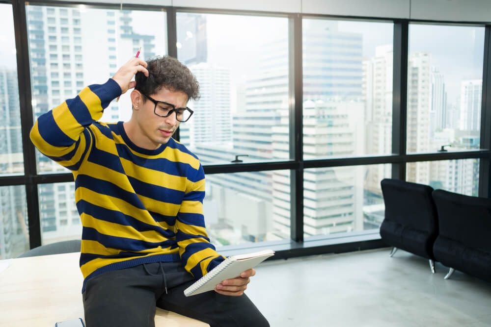 New Young Generation of Businesspeople in a Casual Sweater Is Sitting on the Desk Put His Hand on the Head and Look at the Notebook in Confusion in a Modern Office Surrounded by Glass.