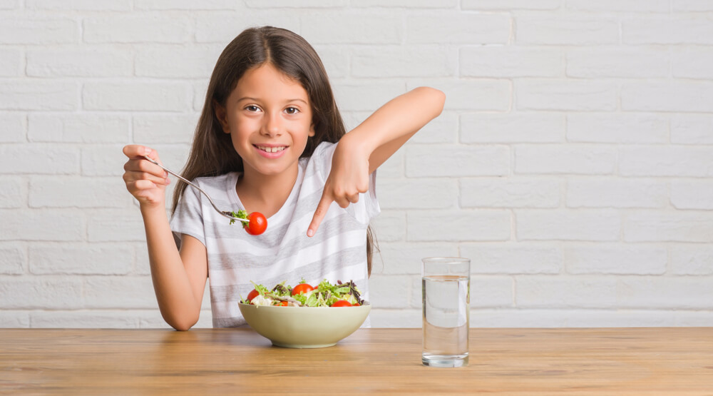 Young Hispanic Kid Sitting on the Table Eating Healthy Salad Very Happy Pointing With Hand and Finger