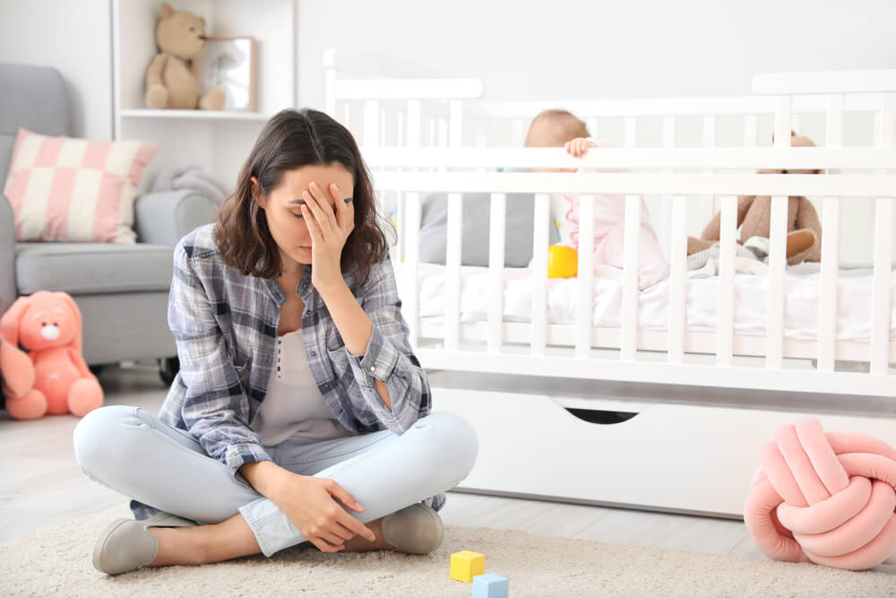 Young Woman Suffering From Postnatal Depression Near Bed With Baby at Home