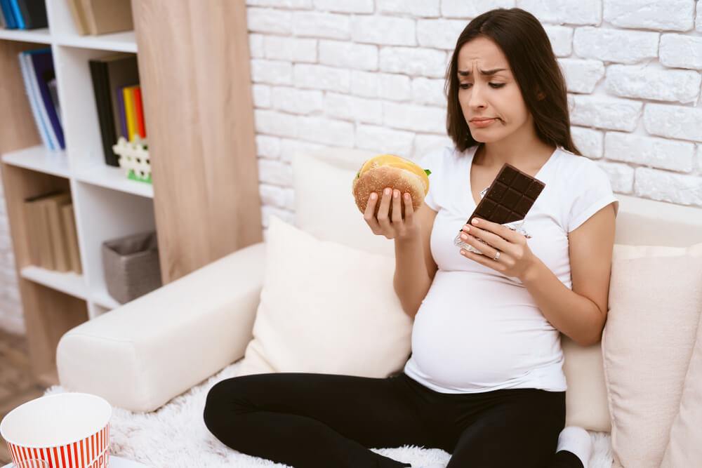Pregnant Girl Eating a Hamburger and Chocolate Sitting on the Couch.
