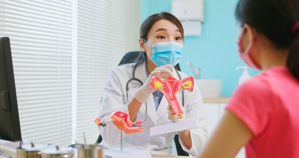 Asian Female Doctor Wear White Coat and Face Mask Explaining Uterus Model to Woman in Hospital