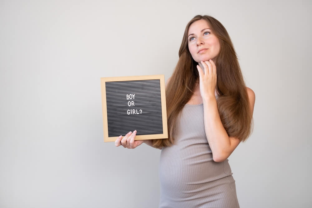 Pregnant Thoughtful Dreaming Woman Holding Board With Boy or Girl Question on Light Gray Background.