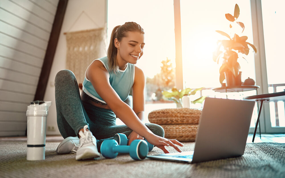 A Sporty Woman in Sportswear Is Sitting on the Floor With Dumbbells and a Protein Shake or a Bottle of Water and Is Using a Laptop at Home in the Living Room