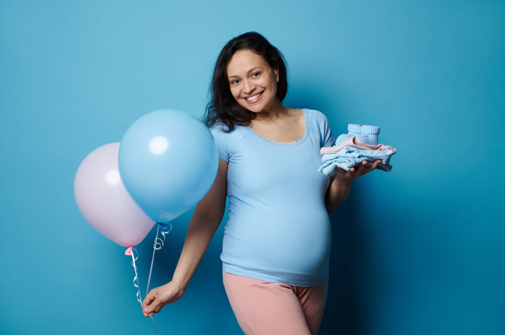 Smiling happy pregnant woman with big belly, holding newborn clothes and two balloons of pink and blue pastel color, isolated on blue background.