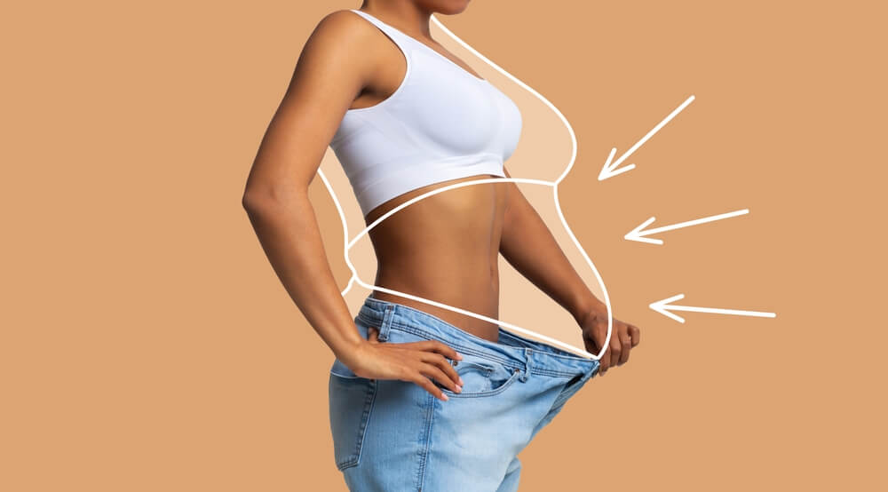 Young slim african american female in big jeans enjoy weight loss result with fat abstract body around, arrows isolated on beige wall background, studio.