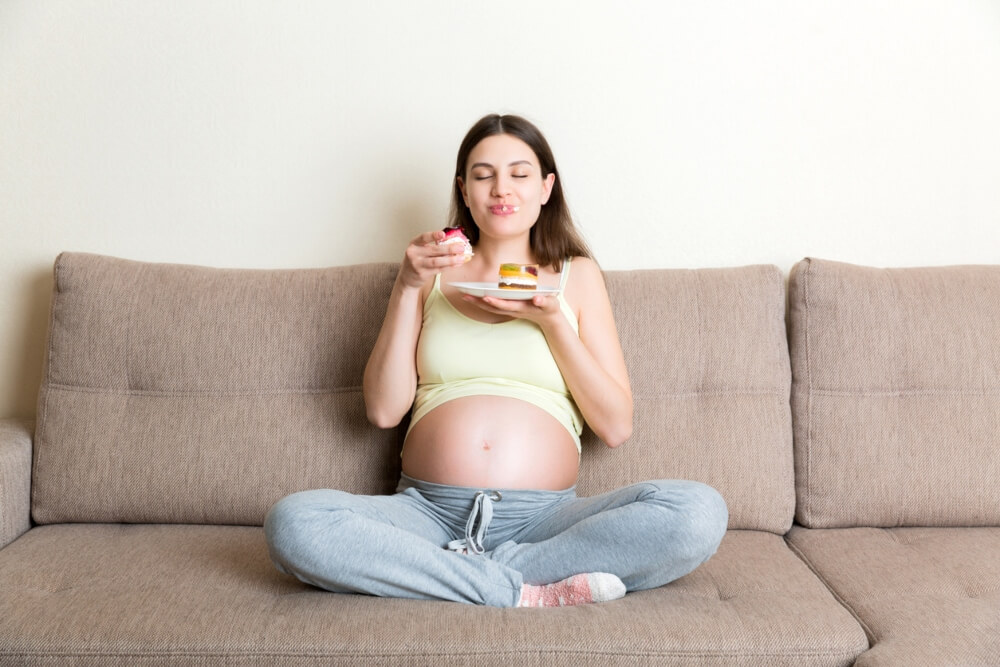 Young Pregnant Woman Enjoys Eating Tasty Cake Resting on the Sofa at Home